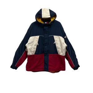 May include: A hooded jacket in navy blue, cream, and red. The jacket features a zip-up front, a hood with a cream stripe, and a small logo on the sleeve. The lower portion of the jacket is red.