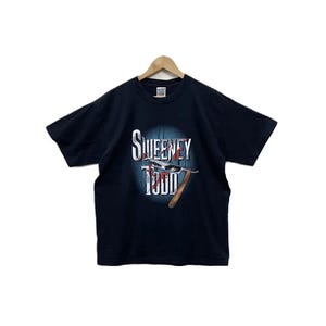 May include: Black t-shirt featuring the text "SWEENEY TODD" in white and red, with a barber razor graphic. The design is set against a blue background. The shirt has a classic crew neck and short sleeves.