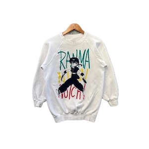 May include: White sweatshirt with a colorful graphic print of a cartoon character from the anime series "Ranna 1/2". The graphic features the character in a fighting pose with the text "RANMA 1/2" written in a stylized font.