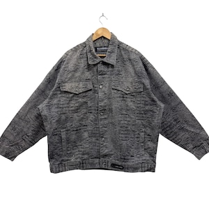 Könnte beinhalten: Eine graue Jeansjacke mit einem strukturierten Muster. Die Jacke hat einen Knopfschließe vorne, zwei Brusttaschen und ein Markenetikett mit der Aufschrift "Pelle Pelle".