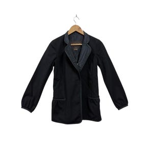 May include: A black jacket with long sleeves and a contrasting gray collar. The jacket has two pockets and a button closure. The jacket is hanging on a wooden hanger against a white background. The jacket appears to be made of a smooth fabric.