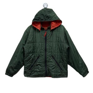 Może przedstawiać: Ciemnozielona pikowana kurtka z kapturem i pełnym zamkiem błyskawicznym. Wnętrze kaptura jest pomarańczowe. Kurtka ma dwie przednie kieszenie i czarny zamek błyskawiczny. Kurtka wisi na białym haczyku.