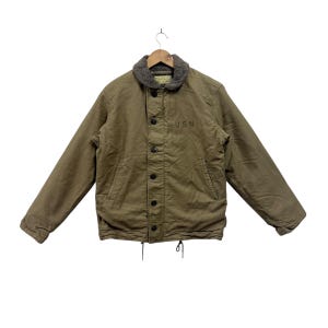 Puede incluir: Chaqueta verde oliva con cuello de piel sintética y cierre de botones. La chaqueta tiene las letras "USN" impresas en la parte delantera. La chaqueta está colgada de una percha de madera.