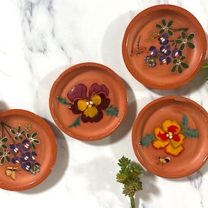 May include: Four small, terracotta-colored dishes with hand-painted floral designs. Each dish features a different flower: purple pansy, yellow and red flower, and two with blue and purple floral arrangements. The dishes are arranged on a white marble surface.