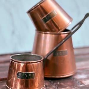 May include: Three copper cider pots with dark handles are stacked on a wooden surface. The front pot has the word "CIDER" on a rectangular label. The pots have a shiny, reflective surface.