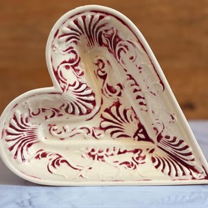 May include: Cream-colored heart-shaped ceramic dish with burgundy floral and scrollwork design. The dish has a raised edge and a shallow interior. The decorative piece is likely used for serving or display.