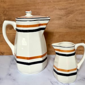 May include: A cream-colored ceramic coffee pot with a lid and a matching creamer pitcher. Both pieces feature a faceted, hexagonal shape and are decorated with horizontal stripes in burnt orange and black around the body.