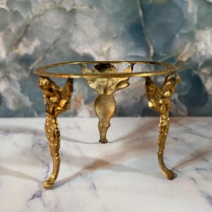 May include: A gold-toned metal display stand featuring a circular top ring supported by three legs shaped like winged figures. The stand has a textured, metallic finish and is designed to hold spherical or round objects securely in place.