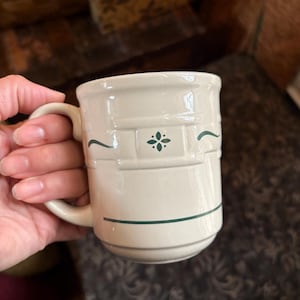 May include: Cream-colored ceramic mug with a handle. The mug features embossed horizontal bands and is decorated with green floral and curved line designs. A classic, simple design for everyday use.