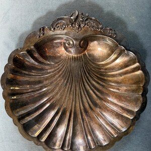 May include: A silver-toned metal dish shaped like a scallop shell. The dish features a deep, fluted interior with radiating ridges and a decorative, ornate scrollwork handle at the top edge. The surface has a polished finish with visible patina.