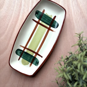 May include: Rectangular ceramic serving dish with a white interior, a brown rim, and a painted design. The design features green and brown brushstrokes. The dish is on a light brown wooden surface.
