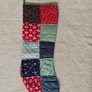 May include: A patchwork Christmas stocking constructed from various fabric squares featuring red, green, blue, and black colors. The patterns include gingerbread men, small hearts, floral prints, and foliage designs. A green fabric loop is attached at the top.