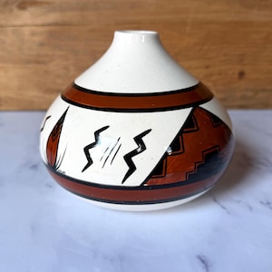 May include: A small, round ceramic vase with a cream-colored base. The vase features horizontal brown bands, black lightning bolt motifs, and geometric step patterns in brown and black encircling the midsection of the vessel.