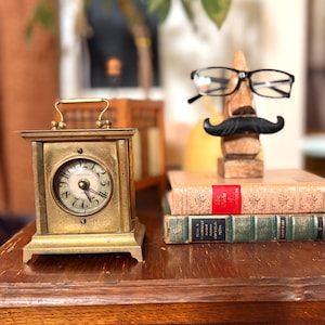 May include: A vintage brass clock with a white face and black numerals sits on a wooden surface. Beside it, a wooden mustache and glasses holder rests on a stack of books. The clock has a handle and a rectangular base.