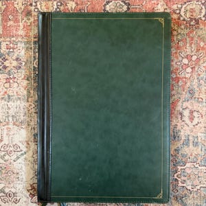 May include: A rectangular dark green portfolio folder with a smooth finish and a black spine. The front cover features a thin gold border with decorative corner accents. A black zipper pull is visible at the bottom left corner of the folder.