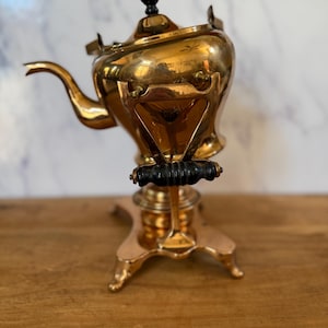 May include: A polished brass tea kettle mounted on a matching brass stand with a curved spout, a black finial on the lid, and a horizontal black handle. The kettle features a rounded body and rests on a four-legged base with a central burner component.
