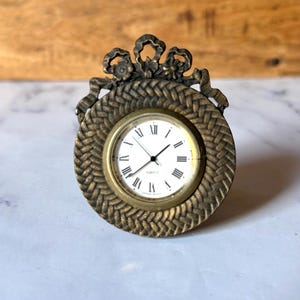 May include: A round quartz clock with a textured, woven-look bronze frame and a decorative bow-shaped top. The clock face is white with black Roman numerals and thin black hands. The piece has a metallic finish and a circular design.