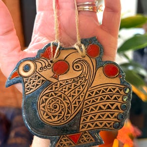 May include: A ceramic bird ornament featuring a blue glazed finish with intricate etched patterns in tan. The design includes vibrant red circular accents on the head, wing, and tail, and is suspended by a thin, light brown jute twine string.