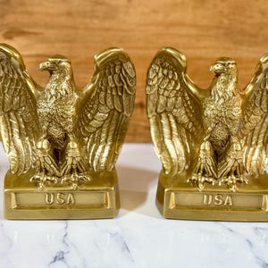 May include: A pair of matching gold-toned metal bookends, each featuring a detailed eagle with wings spread wide. The eagles are perched on a rectangular base that has the letters USA embossed in a clear, raised font on the front face.