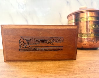 May include: A rectangular wooden box with a carved landscape scene featuring trees, a bridge, and flying birds. The box is a warm brown color and sits on a wooden surface. A copper container with brass accents is in the background.