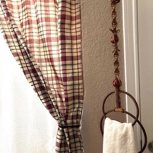 May include: A rustic bathroom scene featuring a plaid curtain in shades of burgundy, cream, and green. A decorative towel holder with a white towel hangs on the wall. The holder is made of metal rings and a decorative rope.