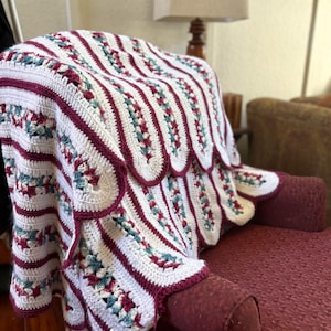 May include: A handmade crochet blanket draped over a red armchair. The blanket features a white base with vertical stripes and scalloped edges in burgundy. Accents of teal and burgundy are incorporated into the design.