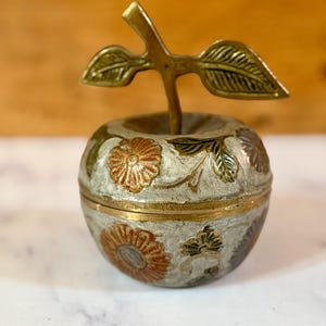 May include: A brass apple-shaped trinket box with a textured silver-toned finish. The surface features embossed floral patterns in copper and green tones. A brass stem with two leaves extends from the top of the lid, which sits on the base.