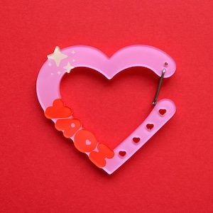 May include: A pink heart-shaped carabiner with the word "AOK" in orange letters. The carabiner has a silver clip and small heart-shaped cutouts. The design includes star accents and is set against a red background.