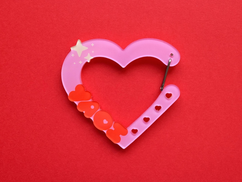 May include: A pink heart-shaped carabiner with the word "AOK" in orange letters. The carabiner has a silver clip and small heart-shaped cutouts. The design includes star accents and is set against a red background.