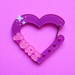 May include: A purple heart-shaped carabiner with the word "URRE" in pink letters. The carabiner has a silver clasp and is decorated with star and heart shapes. The background is a solid pink color.