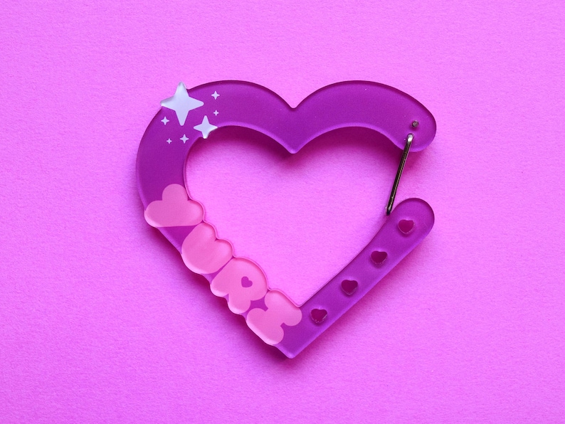 May include: A purple heart-shaped carabiner with the word "URRE" in pink letters. The carabiner has a silver clasp and is decorated with star and heart shapes. The background is a solid pink color.