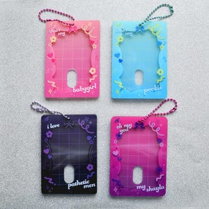 Op de afbeelding: Vier kleurrijke ID-badgehouders met decoratieve frames en zinnen. De houders zijn roze, blauw, paars en magenta, elk met een andere zin: "babygirl", "pookie", "pathetic men" en "my shayla". Elk heeft een zilveren kogelketting.