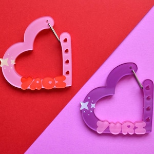 May include: Two heart-shaped earrings. One is pink with the name "YAOE" in red, and the other is purple with the name "YURI" in pink. Each earring has a star design and a silver clasp. The earrings are on a red and pink background.