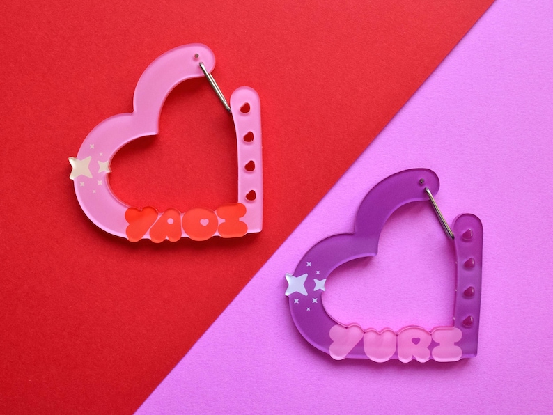 May include: Two heart-shaped earrings. One is pink with the name "YAOE" in red, and the other is purple with the name "YURI" in pink. Each earring has a star design and a silver clasp. The earrings are on a red and pink background.