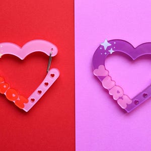 May include: Two heart-shaped carabiner clips. One is pink with the word "YAYOH" in orange, the other is purple with the word "LOVE" in pink. Both have a silver clasp and star accents. The background is split between red and pink.