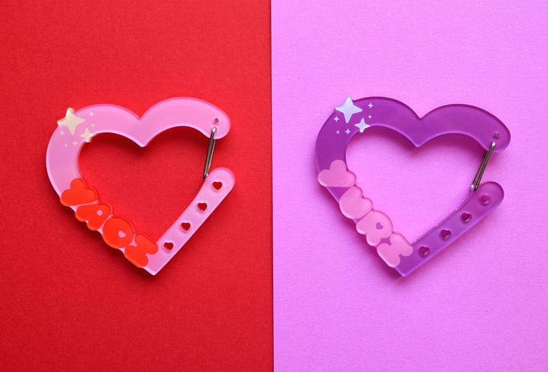 May include: Two heart-shaped carabiner clips. One is pink with the word "YAYOH" in orange, the other is purple with the word "LOVE" in pink. Both have a silver clasp and star accents. The background is split between red and pink.