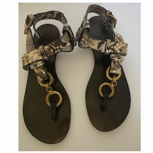 Puede incluir: Un par de sandalias de cuero negro con estampado de piel de serpiente y detalle de cadena dorada.