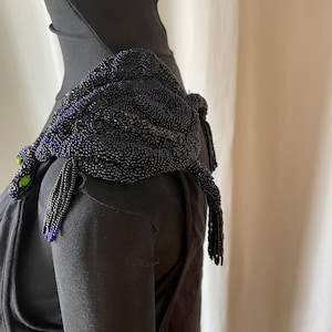 Könnte beinhalten: Ein schwarzes Perlen-Schulterstück in Form eines Drachen mit grünen Augen und violetten Akzenten. Der Drache besteht aus kleinen schwarzen Perlen und hat einen langen Schwanz mit Quasten.