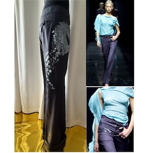 Peut inclure: Pantalon noir avec une large ceinture et un motif d'hippocampe sur le côté. L'hippocampe est fait d'un matériau argenté texturé. L'image montre également un modèle portant un haut bleu clair et un pantalon foncé.