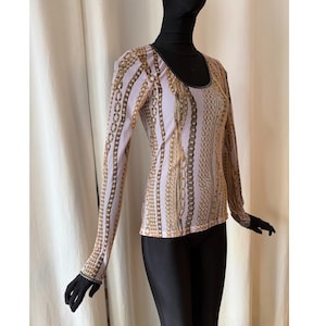 May include: A long-sleeved top featuring a chain design in gold and brown hues against a pale pink backdrop. The scoop-neck top is displayed on a mannequin, with black gloves and trousers.