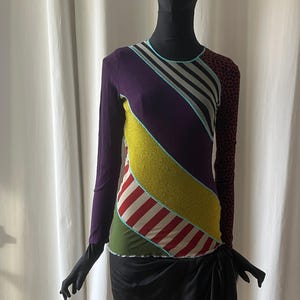 Peut inclure: Un haut à manches longues avec un design patchwork. Le haut présente des sections violettes, jaunes, vertes et des rayures rouges et blanches. La manche droite a un imprimé léopard marron et noir. Le haut est associé à une jupe noire.