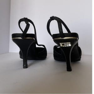 Pode incluir: Um par de saltos pretos com tiras nas costas e saltos agulha. Os saltos têm um detalhe prateado no topo e um pequeno desenho quadrado na parte de trás. Os sapatos estão numa superfície branca.