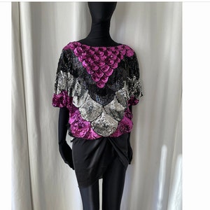 Può includere: Un top di paillettes con un motivo nero, argento e rosa. Il top ha una scollatura rotonda e maniche corte.