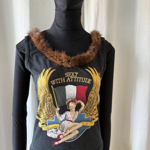 May include: Black tank top with a brown fur collar and a graphic print. The print features a woman, wings, and the text "SEXY WITH ATTITUDE". The top is layered over a black long-sleeve shirt.