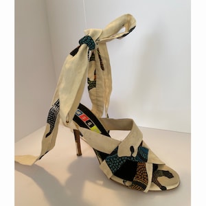 Peut inclure: Sandales à talons blancs avec un tissu à motifs noirs, bleus et jaunes. Les sandales ont une conception de brides croisées et une longue cravate en tissu qui s'enroule autour de la cheville.