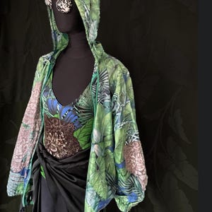 Puede incluir: Una chaqueta con capucha con estampado tropical verde y azul con un traje de baño a juego y una falda negra. La chaqueta tiene cremallera y las mangas son largas. El traje de baño tiene un escote en V profundo. El conjunto se exhibe en un maniquí negro.