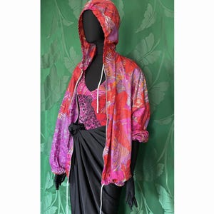 Puede incluir: Una chaqueta con capucha de estampado floral rosa y naranja con cremallera blanca y cordón. La chaqueta se lleva sobre una camiseta negra y una falda negra.