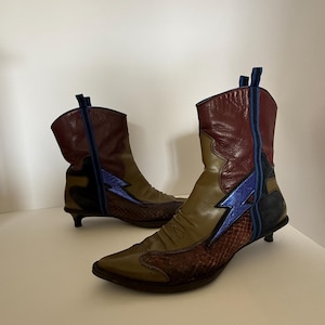 Puede incluir: Un par de botines únicos con un diseño de inspiración occidental. Los botines presentan una mezcla de materiales y colores, incluyendo burdeos, verde oliva y cuero con estampado de serpiente. Un rayo azul adorna el lateral de cada bota. Los botines tienen un tacón pequeño.