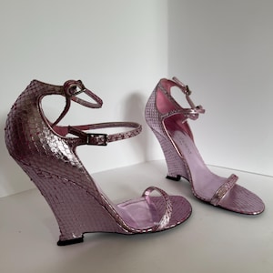 Könnte beinhalten: Ein Paar lavendelfarbene Keilsandalen mit Schlangenmuster. Die Sandalen haben Knöchelriemen und einen einzelnen Riemen über den Zehen. Die Keilabsätze sind geschwungen und die Innenseite ist in einem helleren Lavendelton gehalten.