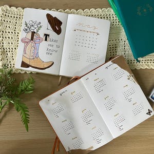 Può includere: Diario aperto con un'illustrazione di stivale da cowboy, un cappello marrone e la scritta "Takes one to know one". Il diario è aperto su una pagina di calendario per maggio. Sono visibili anche altri diari, pastelli e fiori.